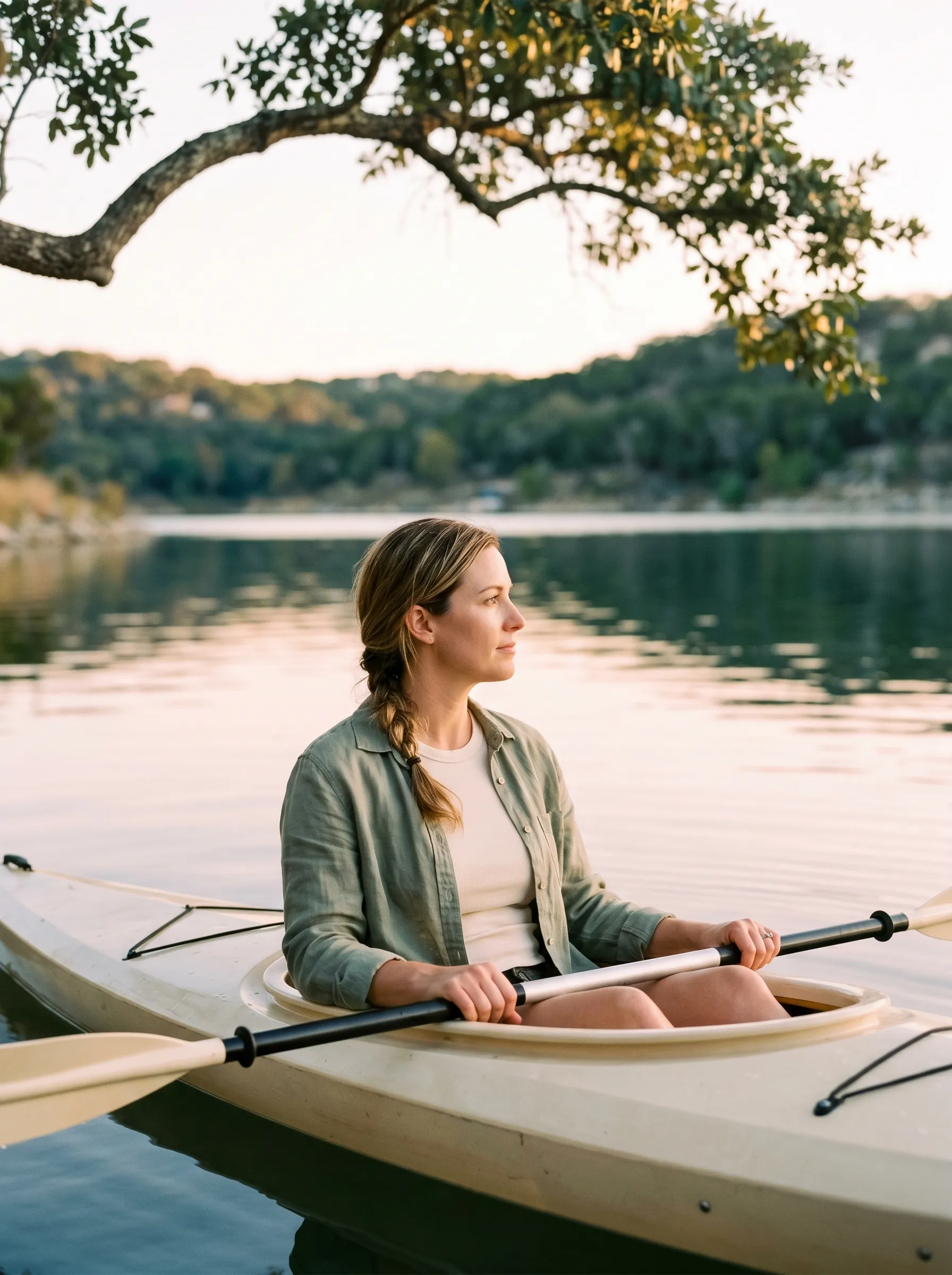 A white woman in her late 30s seated in a simple cream wooden kayak gliding across a calm cove of Lake Travis at warm golden sunrise, a wood paddle resting across her knees, calm grounded gaze across the water.