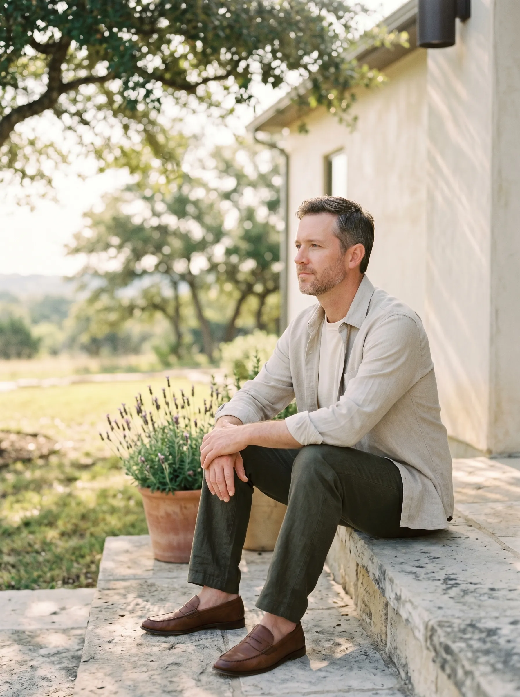 A white man in his early 40s in three-quarter profile seated on a wide weathered limestone front step of a modern Hill Country home, forearms resting on his knees and hands loosely clasped, calm grounded reflective gaze toward soft morning Hill Country light, dappled live-oak shade above.