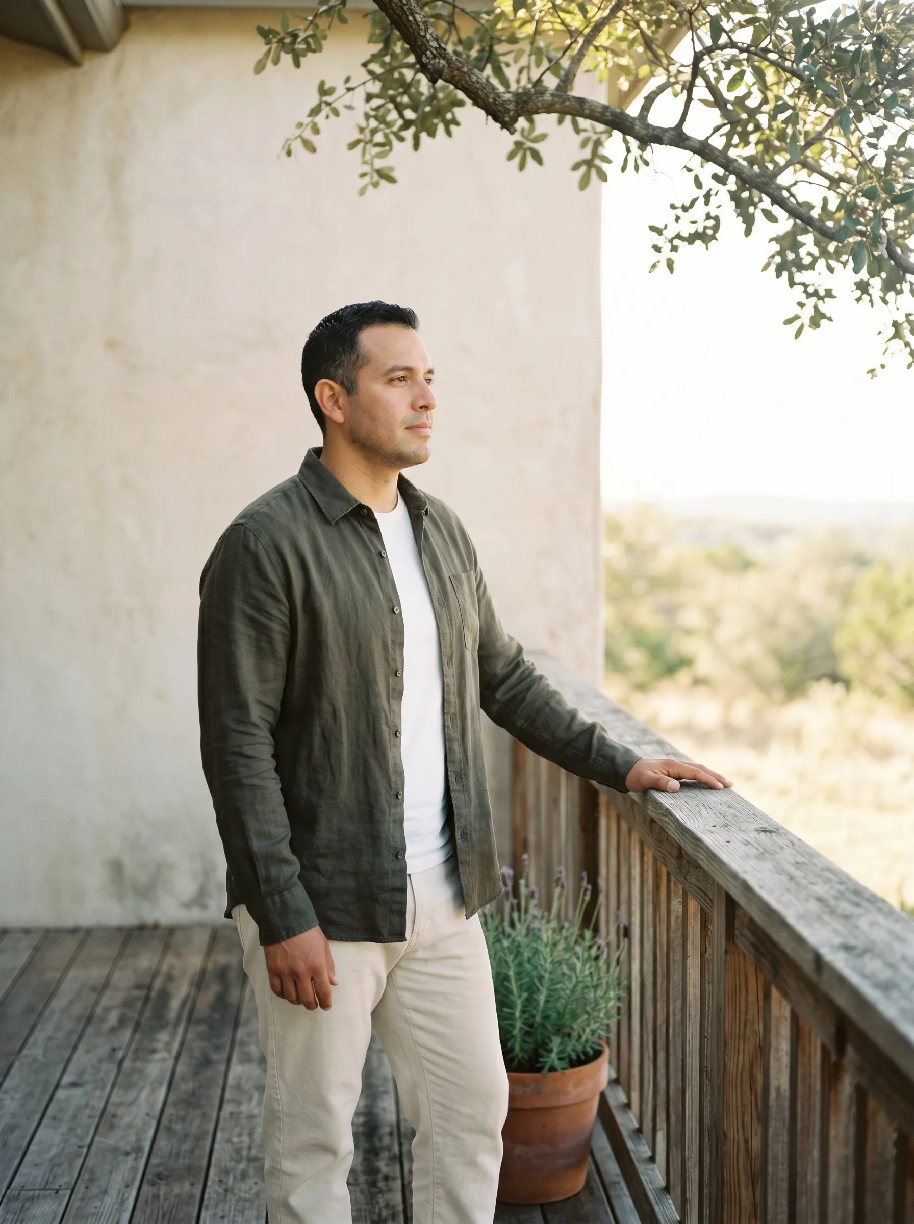 A Hispanic-Latino man in his late 30s standing in three-quarter profile on a covered Hill Country back porch, one hand resting easily on a weathered cedar railing, calm grounded gaze out toward a soft live-oak horizon in late-morning light.