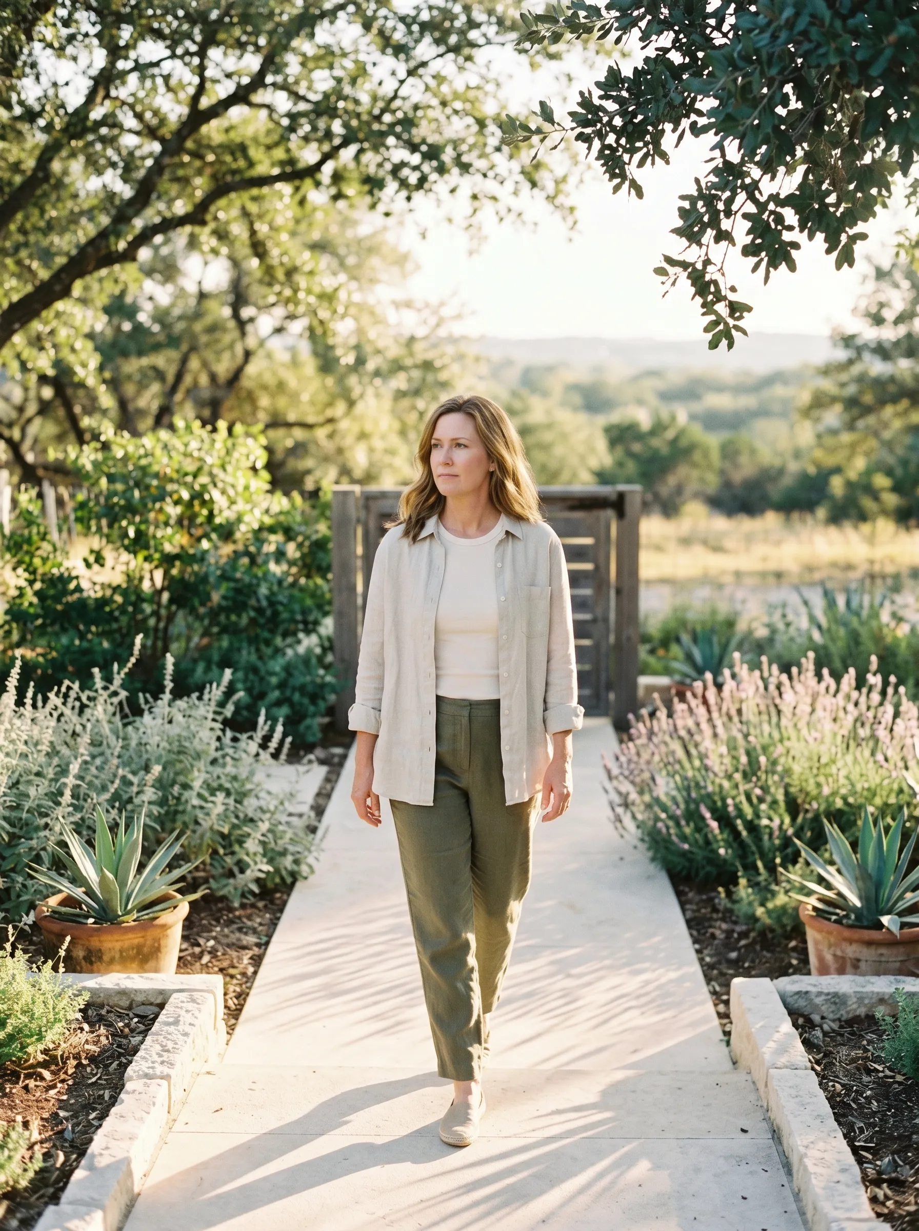 A woman in her late 40s with shoulder-length warm dark blonde hair worn loose, walking forward in three-quarter profile up a smooth limestone path between native silver salvia and agave in a modern Hill Country side yard, calm grounded gaze ahead, late golden afternoon light.