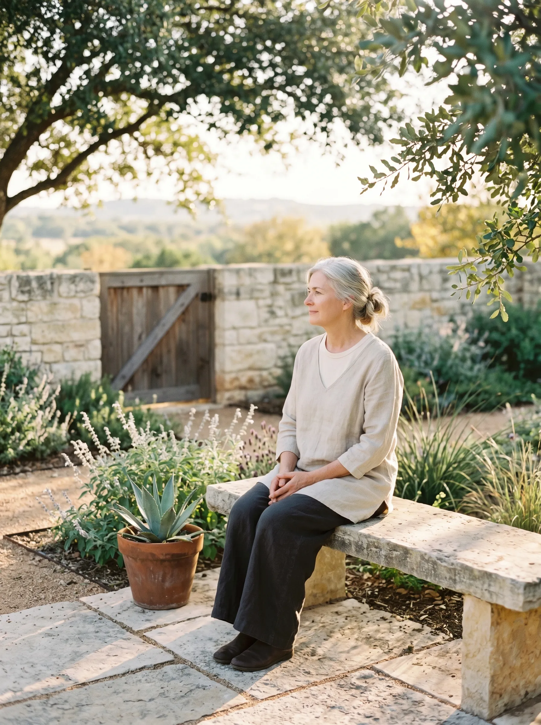 A woman in her late 50s with silver hair in a soft low bun, oat-linen tunic and dark wide-leg linen trousers, seated three-quarter profile on a sun-warmed weathered limestone garden bench in a Hill Country side garden, hands folded gently in her lap, calm grounded reflective gaze toward the soft middle distance, terracotta-potted agave at the bench edge, native silver salvia and lavender in nearby beds, weathered cedar gate behind, late golden afternoon light.