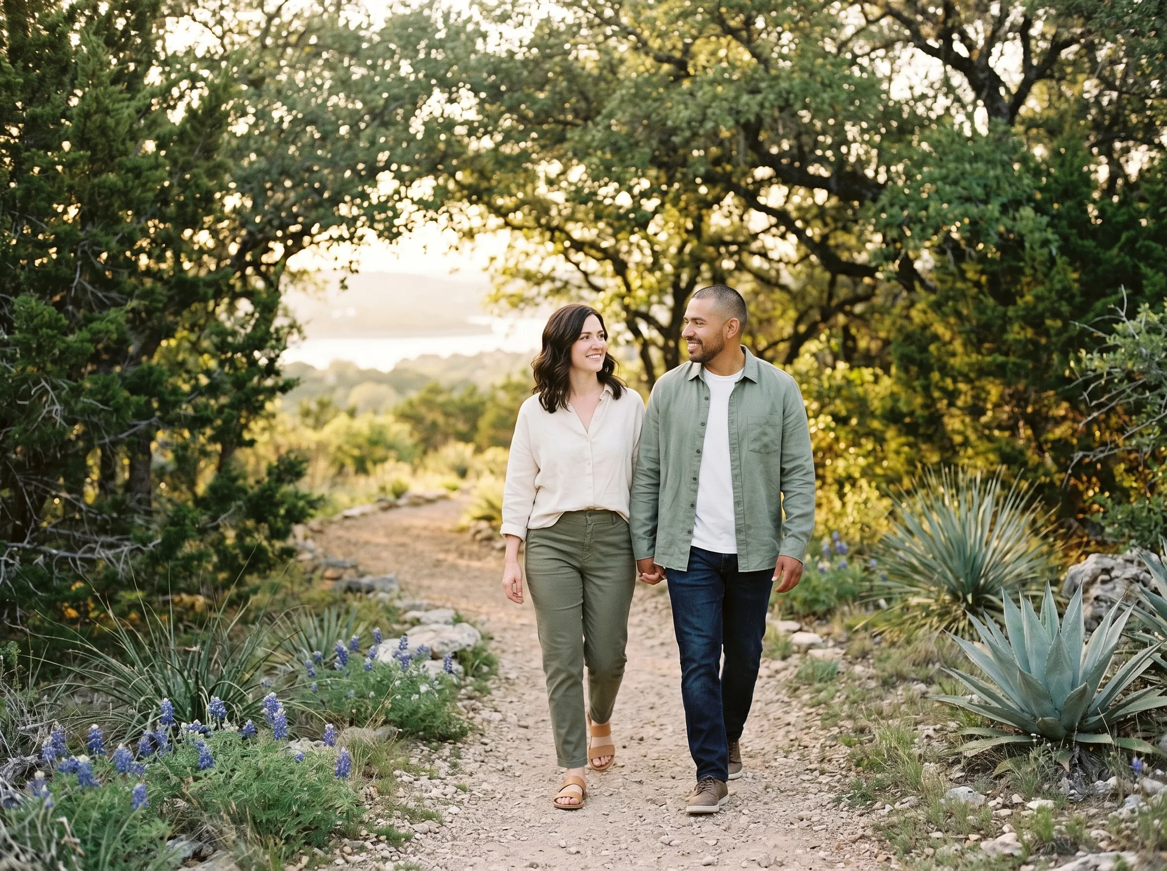 An interracial couple in their mid-30s walking together along a juniper-edged Hill Country path at late golden afternoon, hands close in companionable proximity, Lake Travis softly bokeh in the distance.