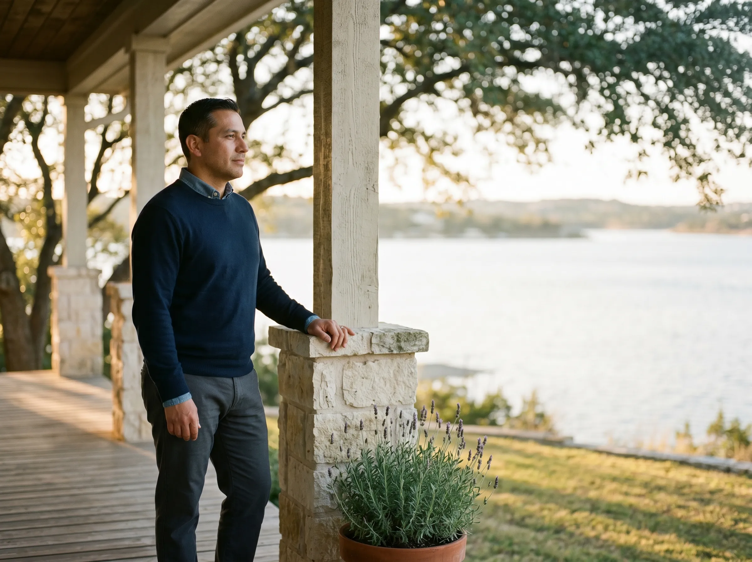 A Hispanic man in his mid-40s on a covered limestone porch overlooking Lake Travis at late golden afternoon, one hand resting lightly on a limestone column, calm grounded gaze across the water.