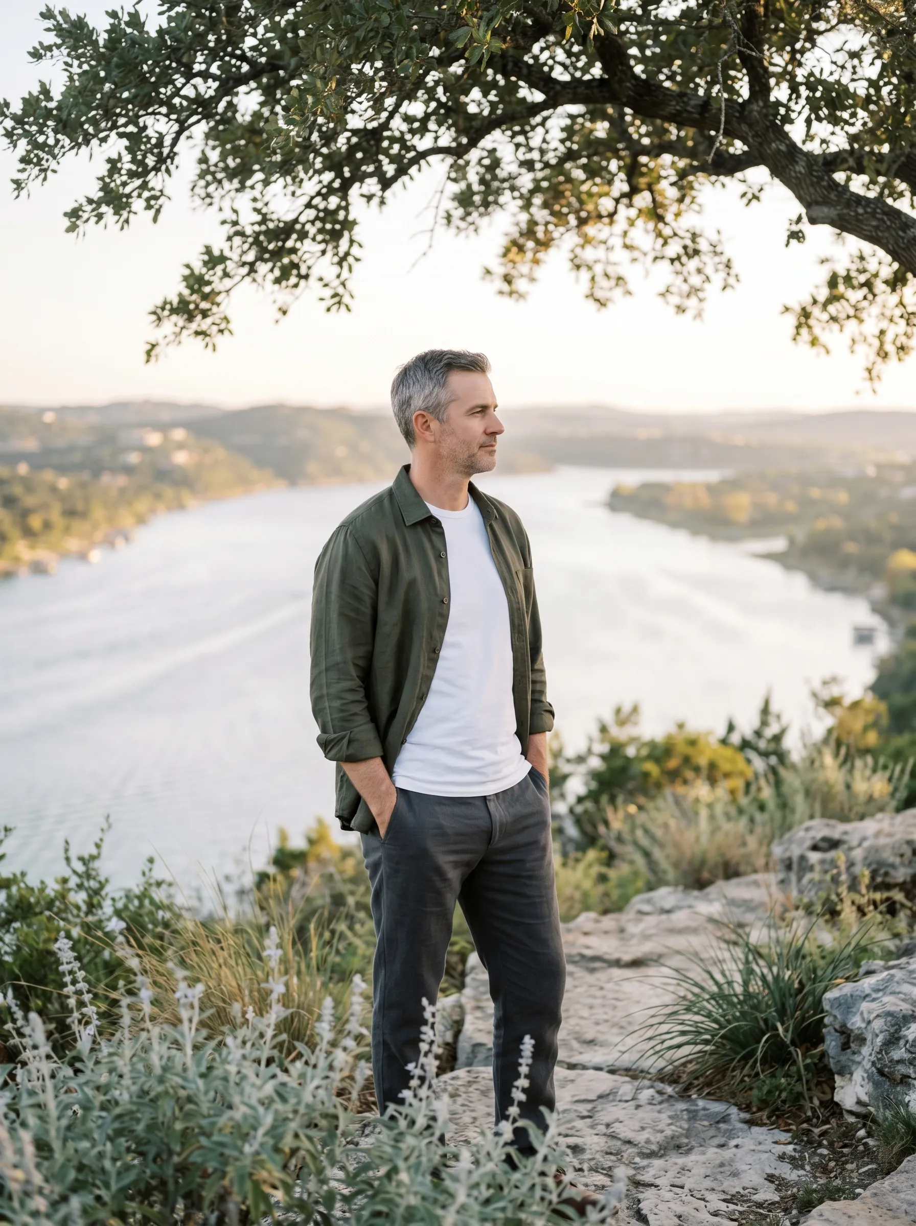 A man in his late 40s standing at a Hill Country limestone overlook at late golden afternoon, both hands resting lightly in his trouser pockets, calm grounded gaze toward Lake Travis spread across the middle distance below.