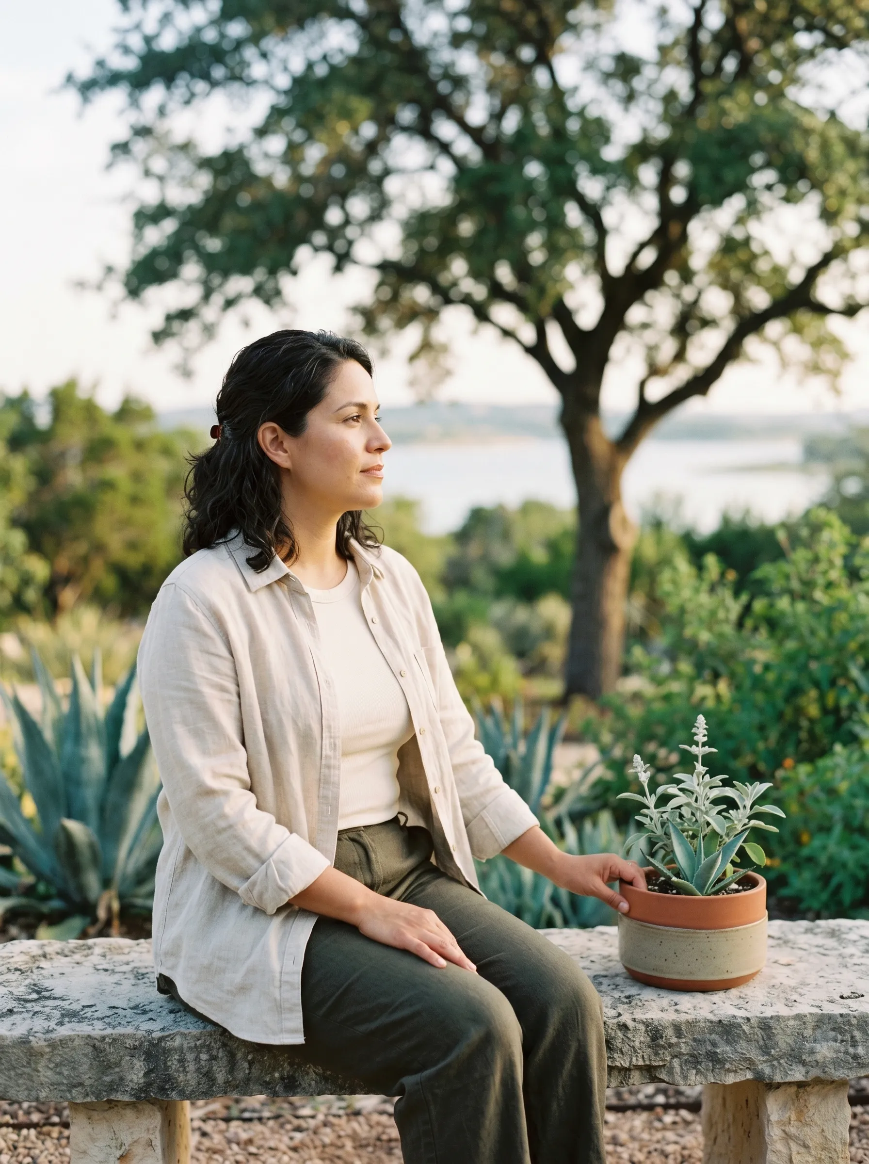 A Hispanic-Latina woman in her late 30s seated three-quarter profile on a sun-warmed limestone bench in a modern Hill Country native plant garden, one hand resting on a stoneware planter of silver salvia, calm grounded reflective gaze toward a single live-oak in the middle distance, agave and pale lake water in soft bokeh beyond, late golden afternoon light.