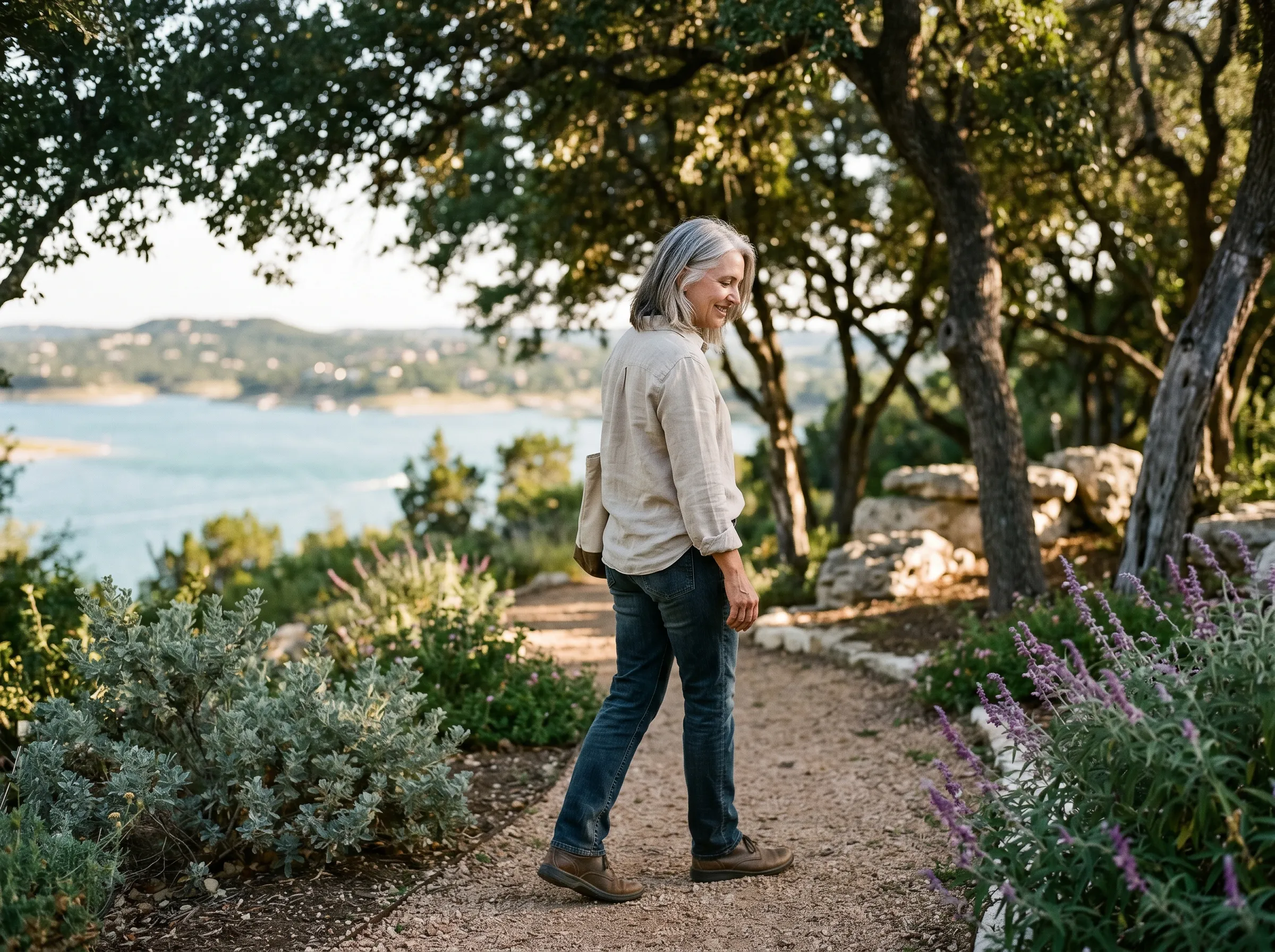A woman in mid-50s walking a quiet path beside Lake Travis in middle-distance bokeh, late afternoon Hill Country light.