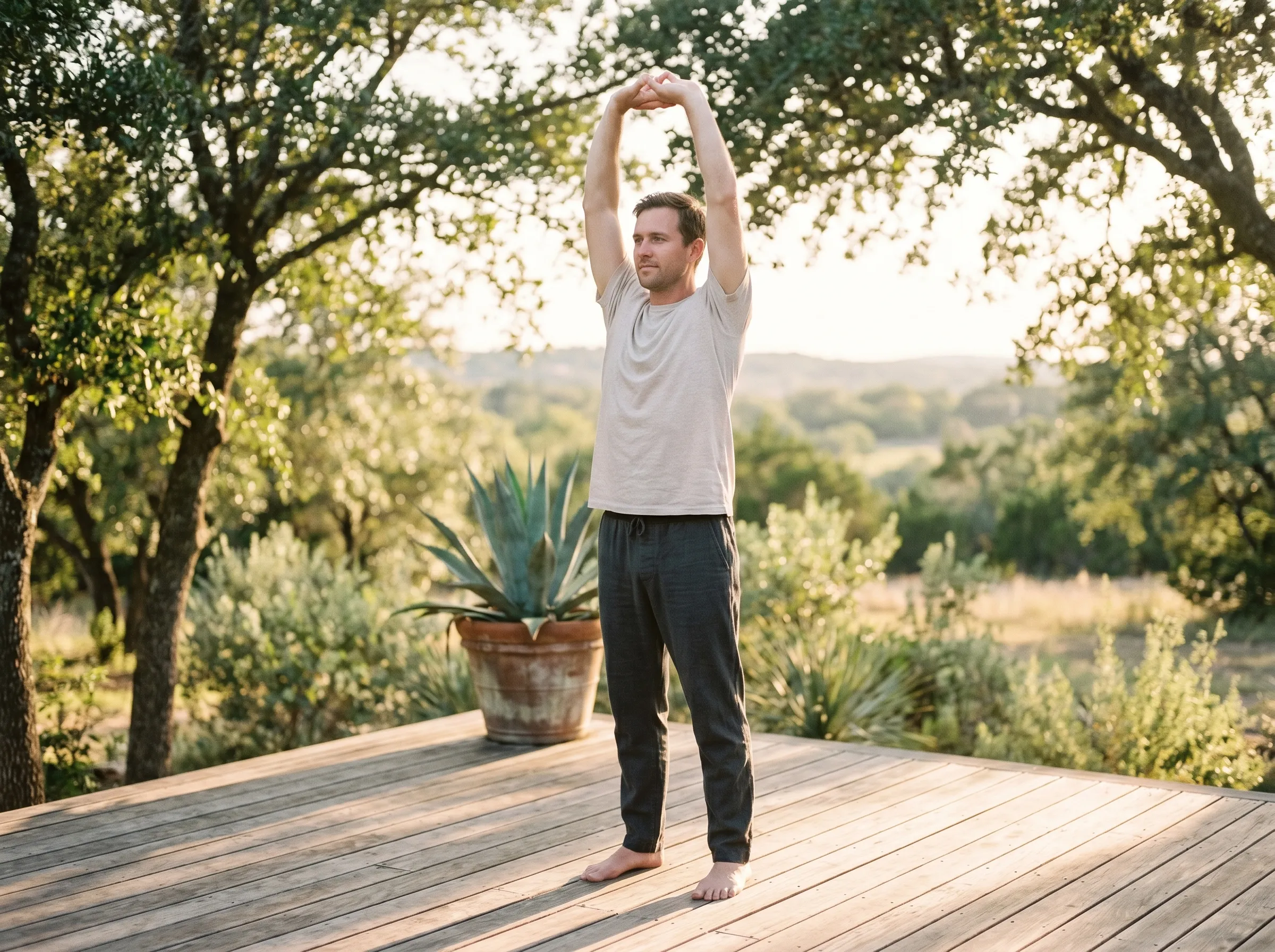 A man gently stretching his arms overhead on a sunlit cedar deck at golden hour, Hill Country canopy beyond.
