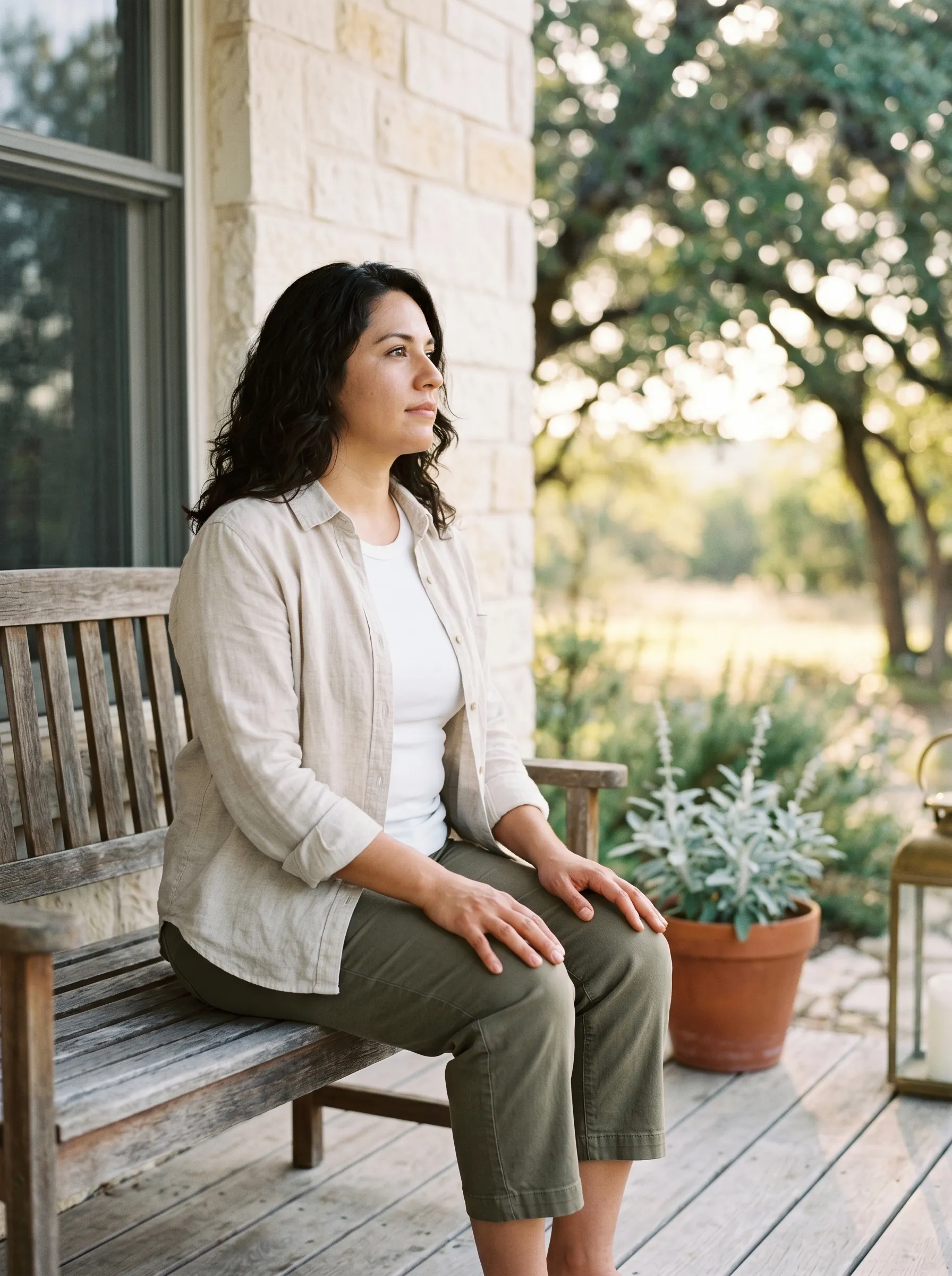 A Hispanic woman in her late 30s seated on a weathered cedar bench on a covered limestone porch of a modern Hill Country home, three-quarter profile, hands resting open on her knees, calm grounded gaze toward dappled late golden afternoon light through live oak.