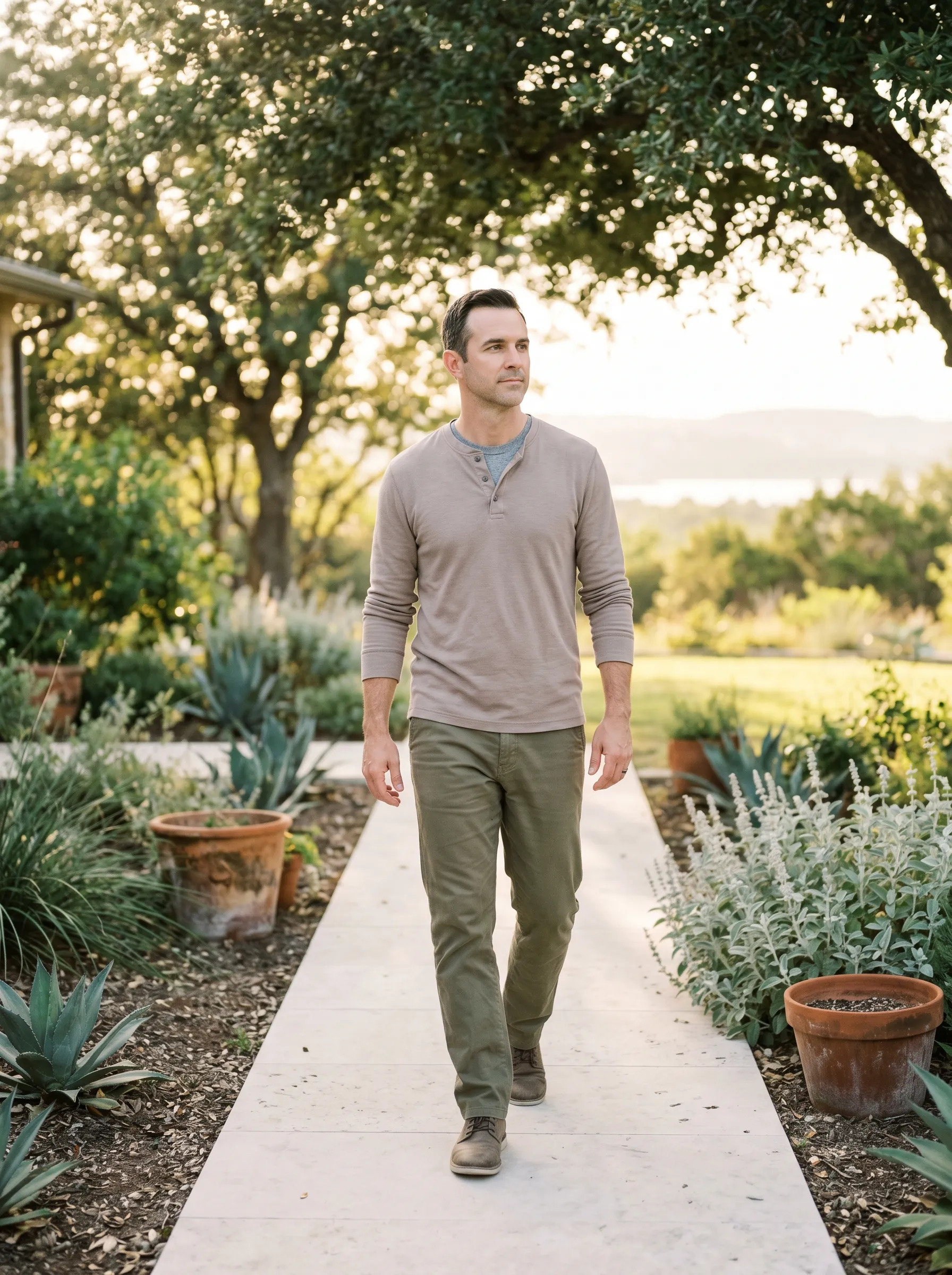 A white man in his early 40s walking forward along a smooth limestone garden path between agave and silver salvia, full body three-quarter profile, calm grounded expression, dappled live-oak shade, warm late golden afternoon Hill Country light.