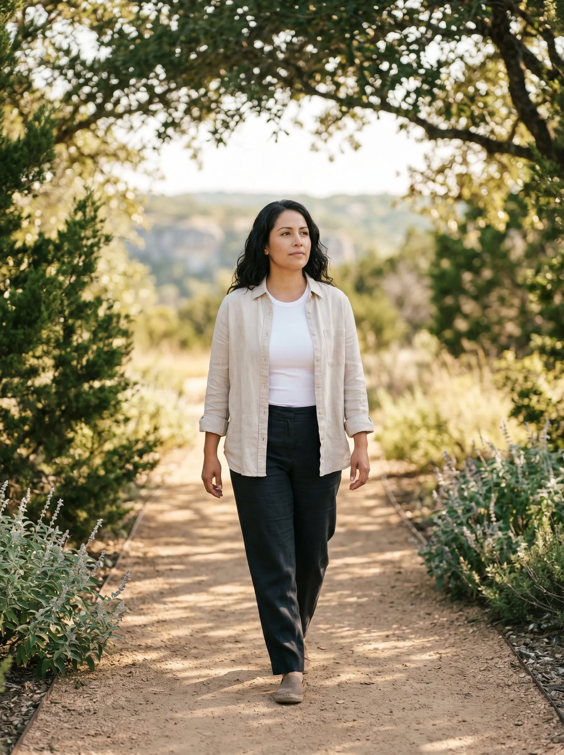 A Hispanic-Latina woman in her late 30s walking forward in three-quarter profile on a juniper-edged Hill Country trail, oat-linen overshirt over white tee and dark linen trousers, dappled live-oak canopy and a distant limestone bluff softening to gentle bokeh in late-morning light.
