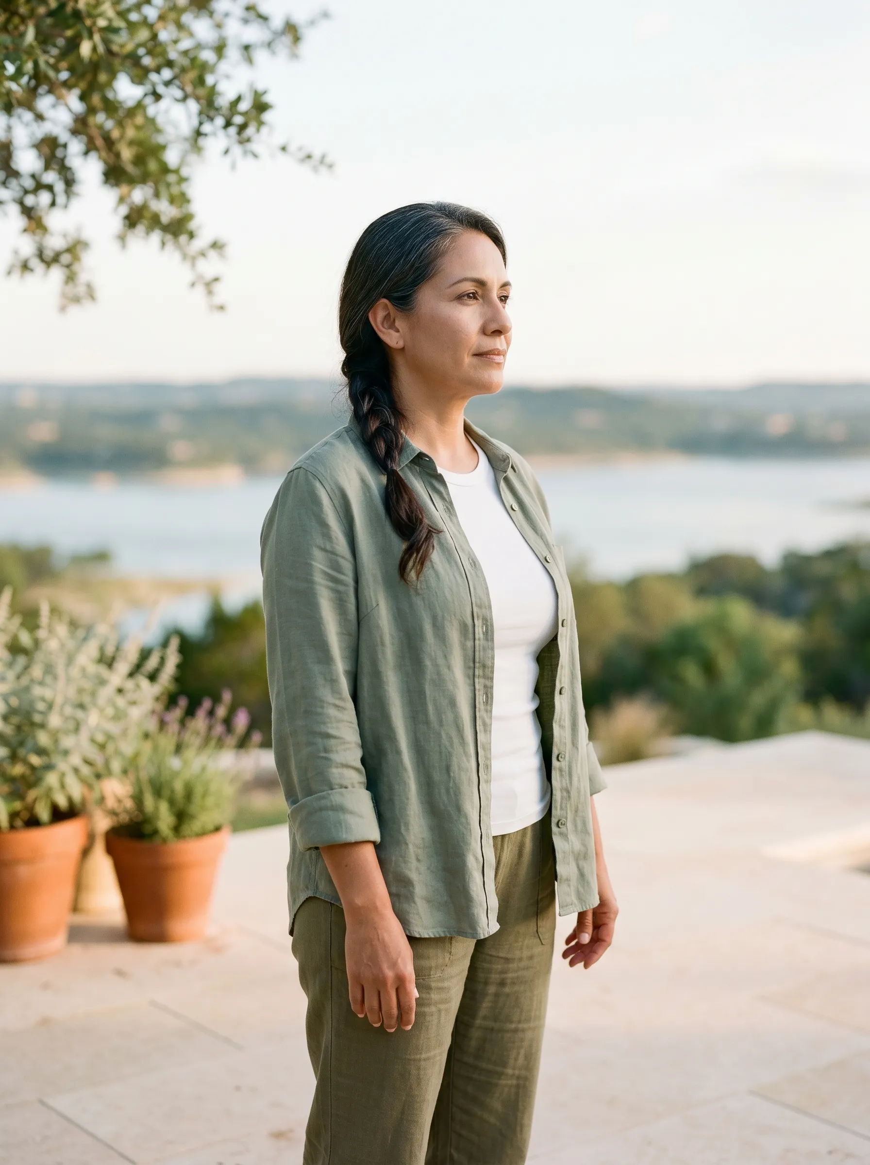 A Hispanic-Latina woman in her mid-40s standing in three-quarter profile on a sun-warmed limestone patio at the edge of a Lake Travis vista, hands relaxed at her sides, calm grounded gaze toward distant water, late golden afternoon Hill Country light.
