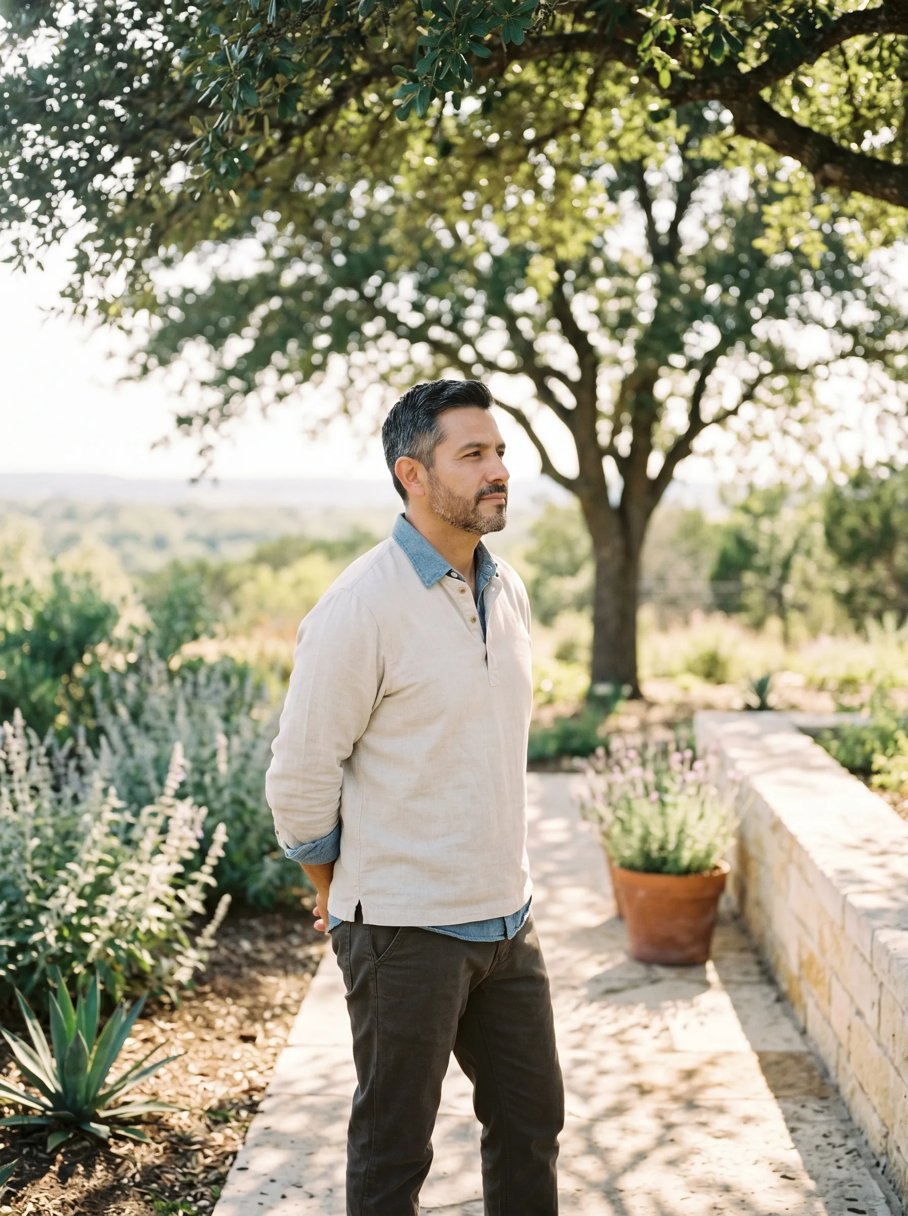 A Hispanic-Latino man in his late 40s standing three-quarter profile on a sun-warmed limestone garden path in a modern Hill Country native plant garden, hands clasped loosely behind his back, calm grounded reflective gaze toward a single live-oak in the middle distance, agave and silver salvia framing the path, late golden afternoon light.