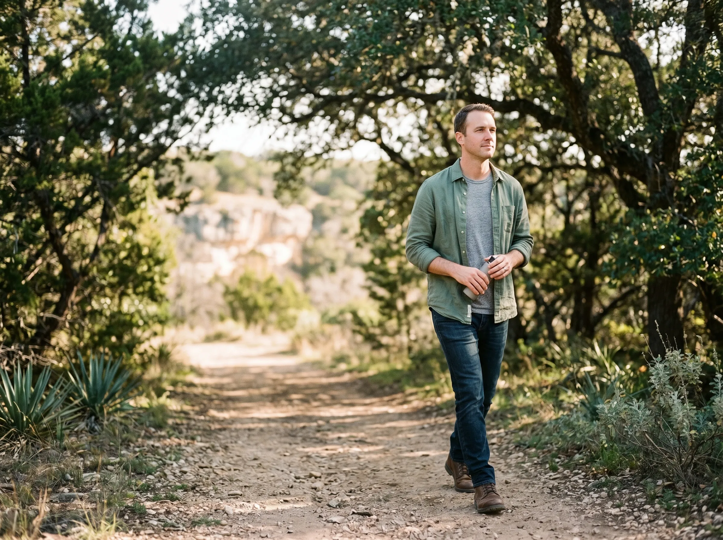 A man in his late 30s walking a juniper-edged Hill Country trail at mid-morning, dappled live-oak canopy overhead.