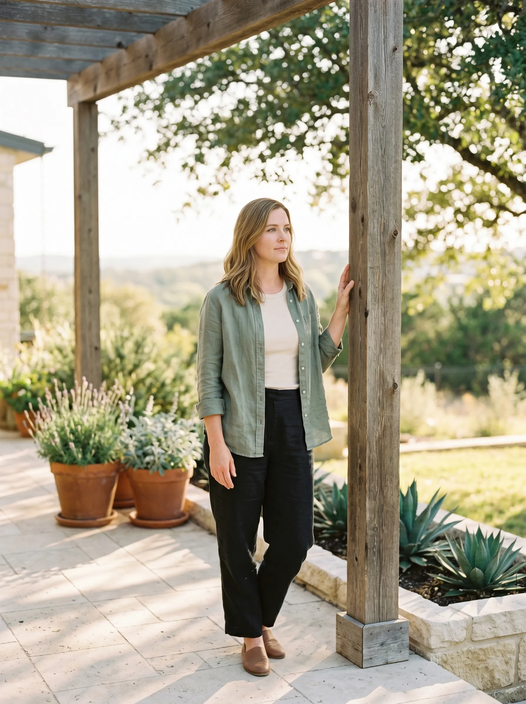 A white woman, late 30s, on a sun-warmed limestone patio of a modern Hill Country home in Bee Cave at warm late golden afternoon, leaning lightly against a weathered cedar pergola post with one hand on the post, terracotta-potted Hill Country lavender along the patio edge and dappled live-oak shade above.