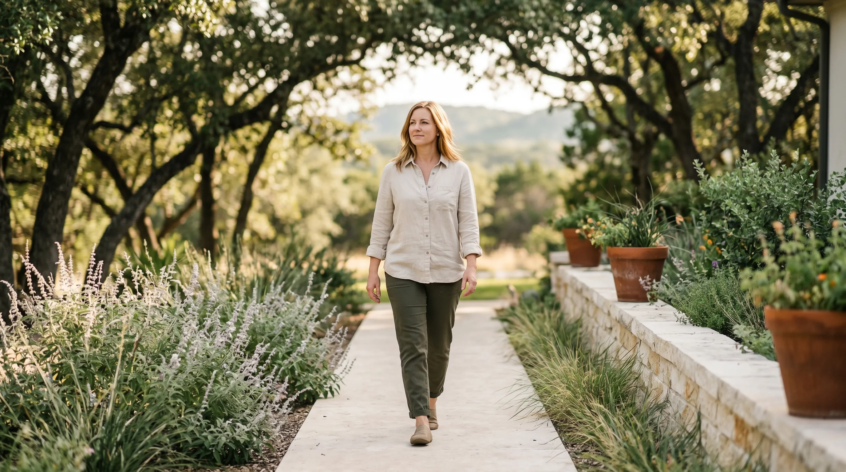 A Hispanic Latina woman in her late 30s walking forward at an easy pace along a smooth limestone path through a wooded Hill Country front yard in Dripping Springs at warm late golden afternoon, sage and silver salvia along the path edges, dappled live-oak canopy shade across the path, distant rolling Hays County hills softly visible in middle-distance bokeh.