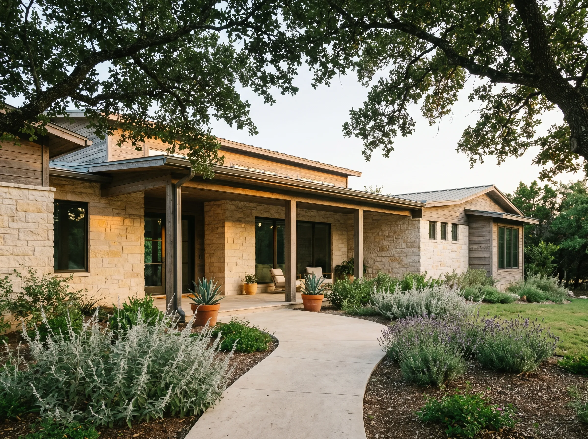 A modern Hill Country home in Lakeway framed by live-oak canopy at warm golden hour, with sun-warmed limestone walls, weathered cedar trim, and a smooth limestone path edged in silver salvia and Hill Country lavender.