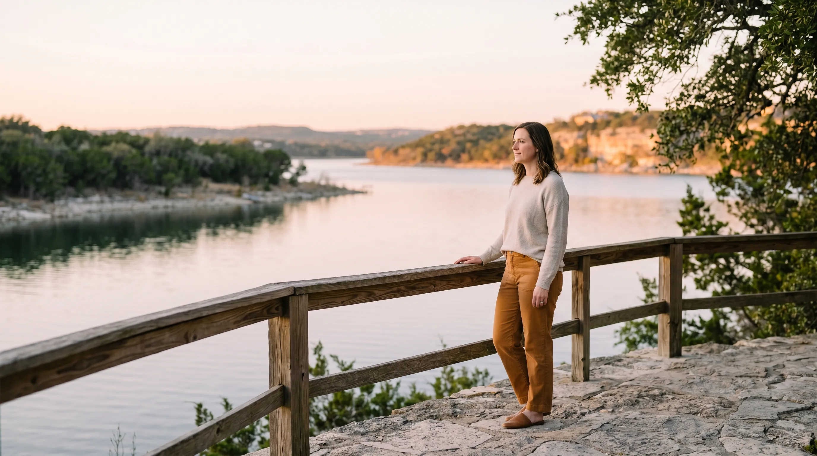 A white woman in her mid-30s in mid-body three-quarter profile on a wide low-limestone overlook above a calm Lake Travis cove at warm late golden afternoon, linen oatmeal sweater and warm caramel trousers, one hand resting on a weathered cedar railing, glassy lake water in the middle distance and sun-warmed limestone bluffs across the cove.