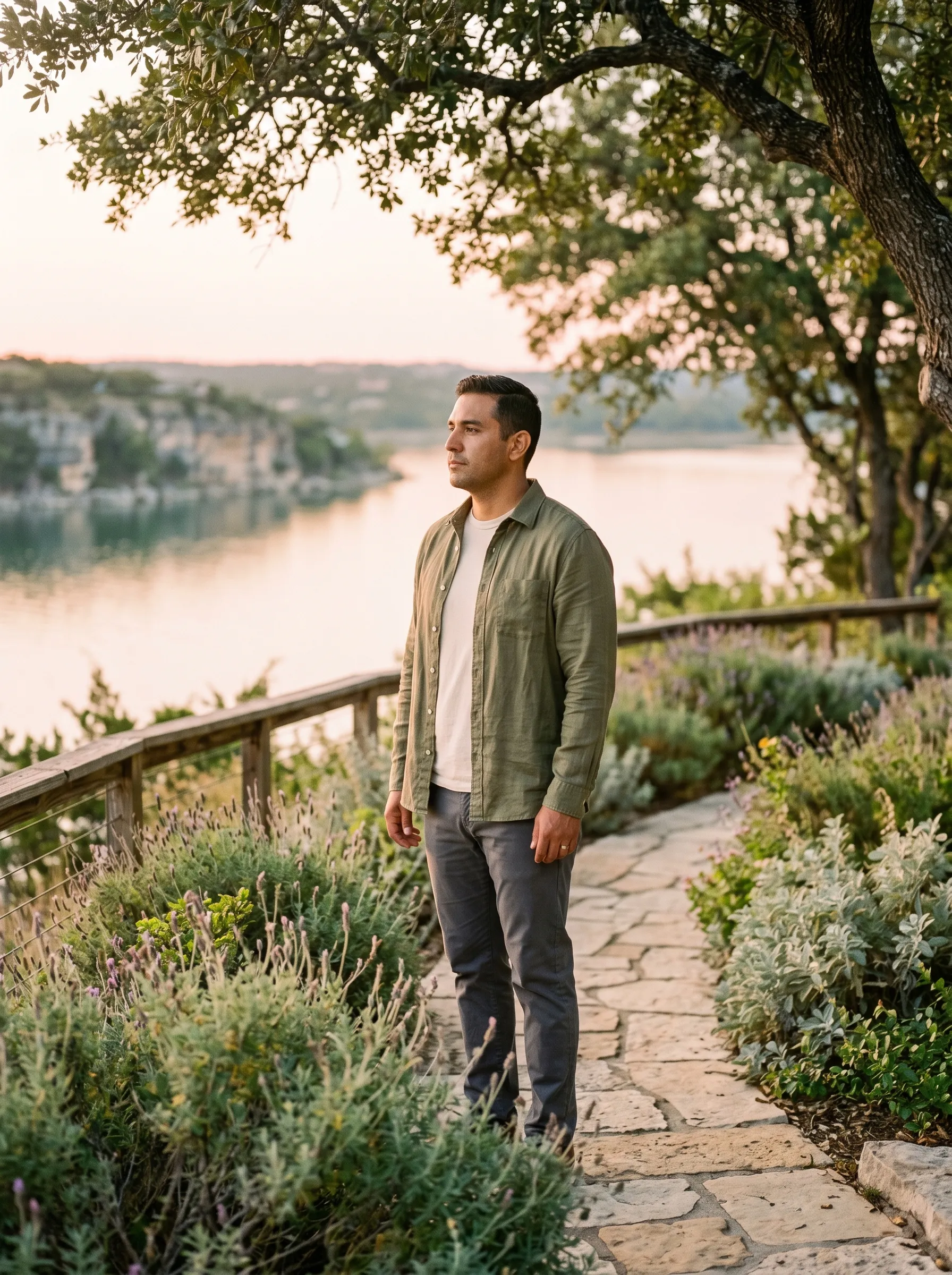 A Hispanic man in his late 30s in mid-body three-quarter profile on a sun-warmed limestone path along a low Lake Travis overlook at warm late golden afternoon, sage linen overshirt over a soft cream tee with charcoal trousers, native Hill Country lavender and silver salvia along the path, a glassy Lake Travis cove with sun-warmed limestone bluffs in the soft middle-distance bokeh, dappled live-oak canopy at the upper edge of the frame.