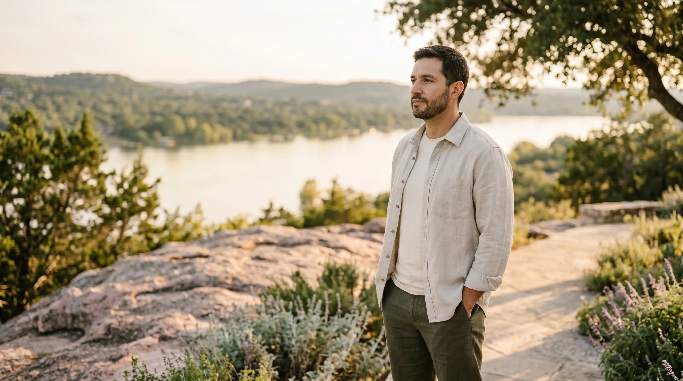 A Hispanic Latino man, late 30s, standing on a sun-warmed granite overlook above Lake Marble Falls in Burnet County at warm late golden afternoon, hands relaxed in linen trouser pockets, calm grounded gaze across the Highland Lakes water and distant Hill Country ridges.