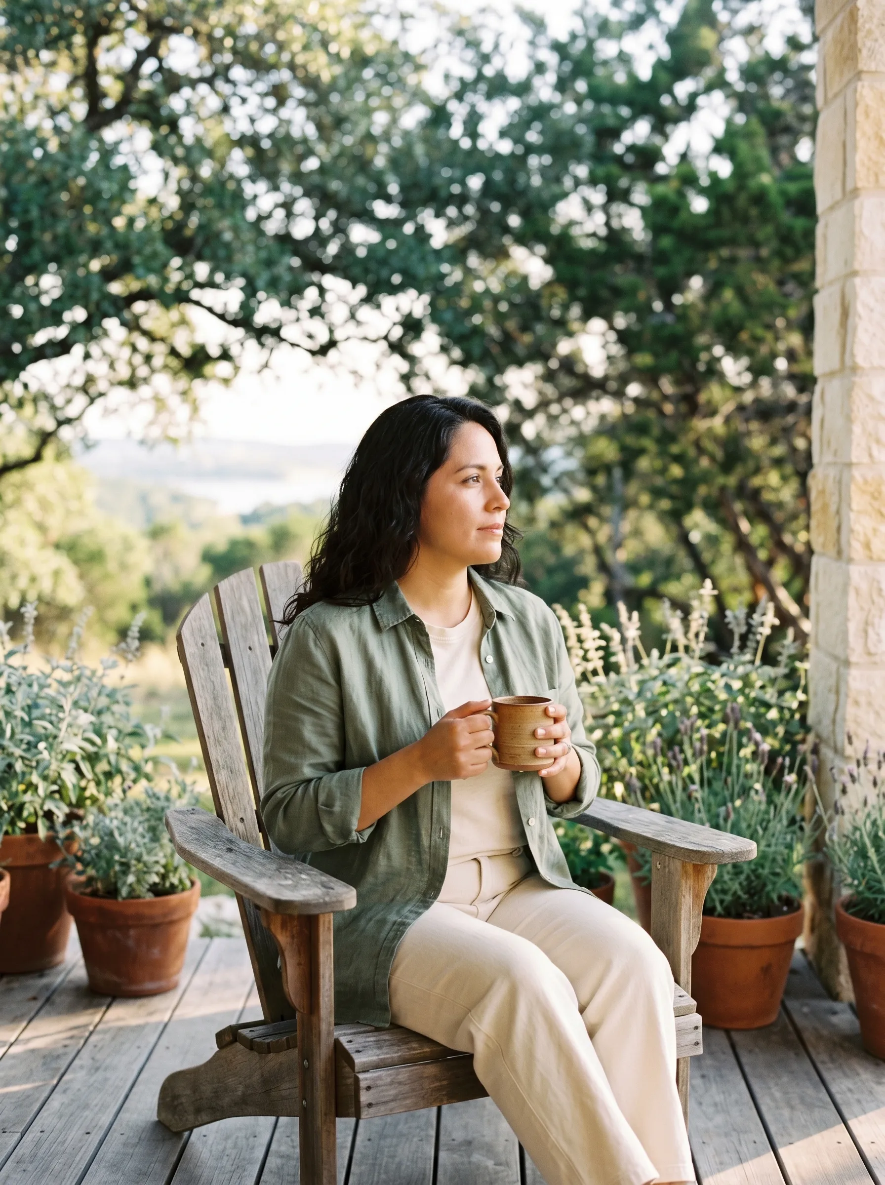 A Hispanic Latina woman in her late 30s with dark wavy shoulder-length hair, sage linen button-up and cream tee, seated relaxed on a weathered wooden Adirondack chair on the covered side patio of a wooded Hill Country home in Spicewood, holding a stoneware mug at chest height, terracotta-potted lavender flanking the patio, dappled live-oak canopy and a distant Hill Country slope into Lake Travis behind.