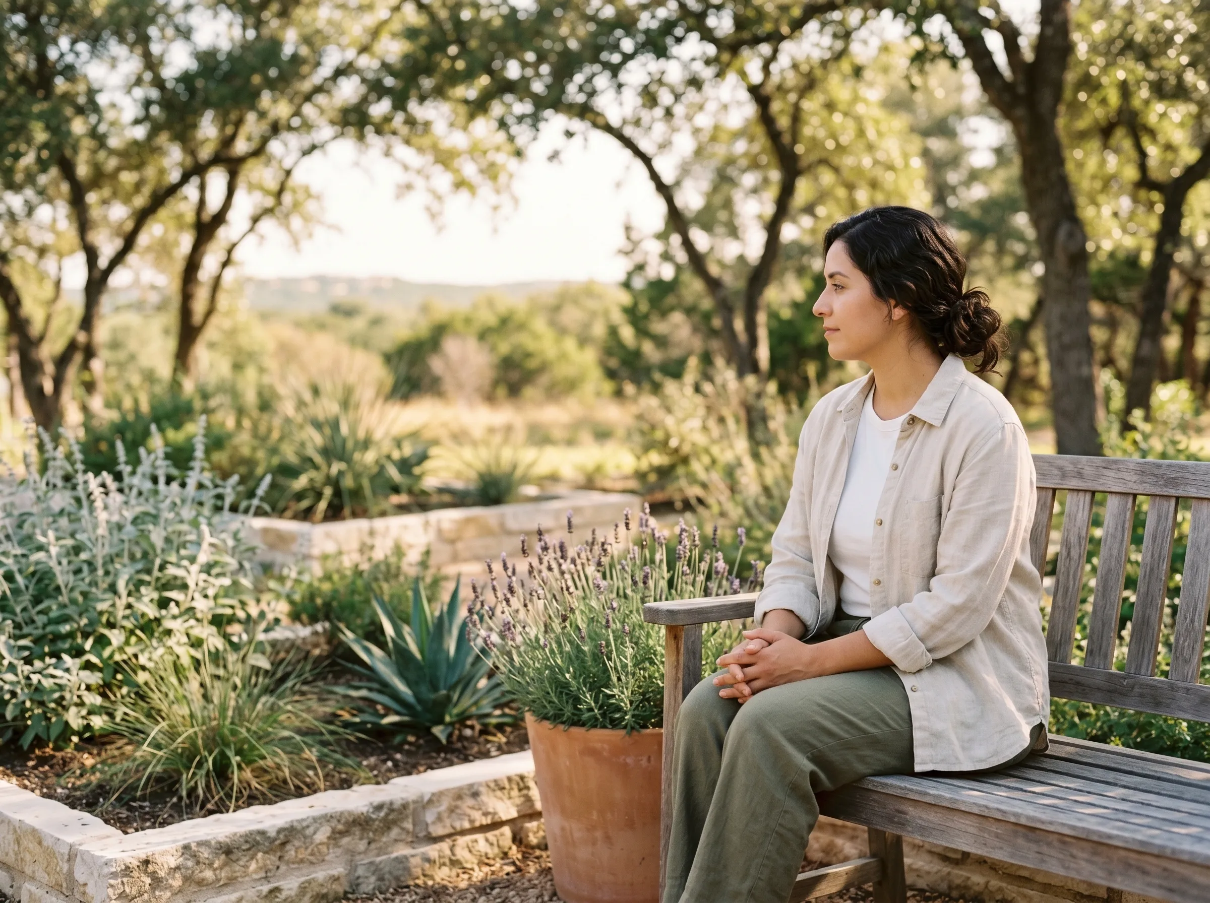 A woman with dark wavy hair seated on a weathered cedar bench in a Hill Country backyard, hands folded gently in her lap, gazing across a quiet native garden of lavender and silver salvia in warm late-morning light.