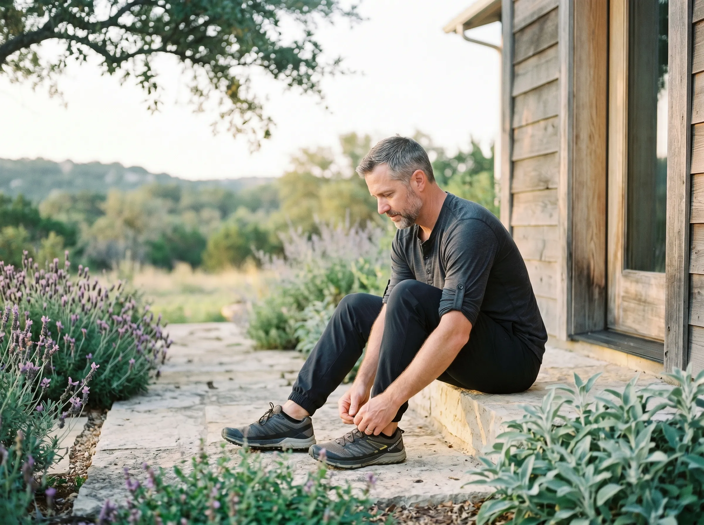 A man with salt-and-pepper hair seated on a sun-warmed limestone front step of a modern Hill Country home, tying his trail running shoe in calm early-morning light, native lavender along the path.