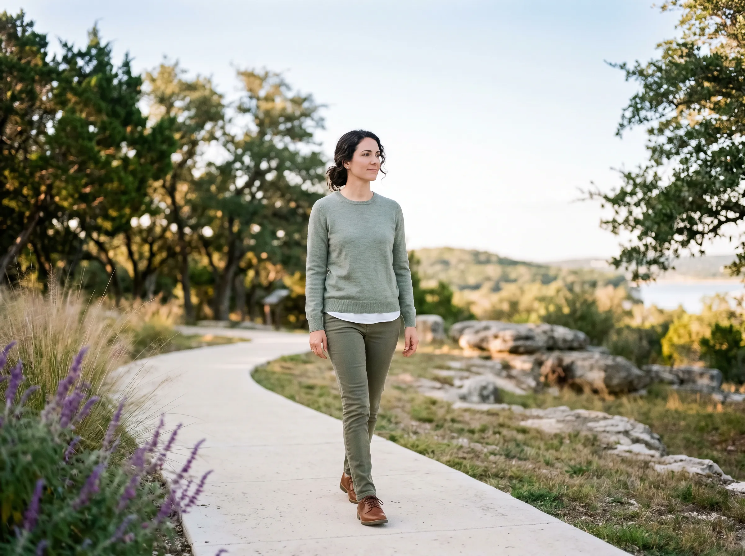A woman walking a Hill Country trail at golden hour, Lake Travis softly visible in the distance.
