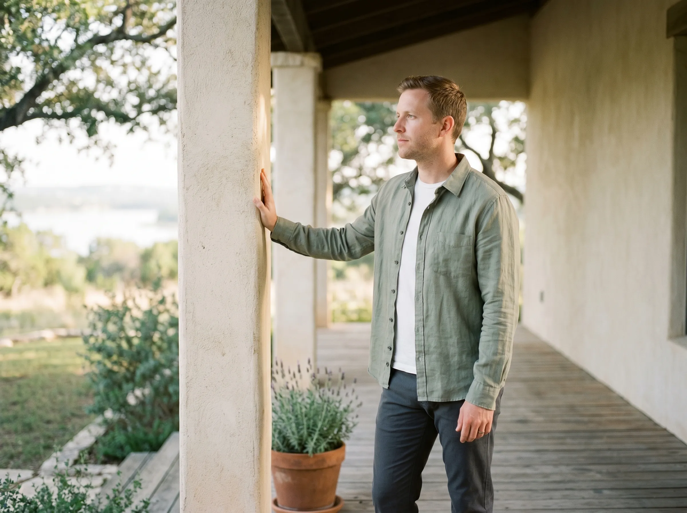 White man in his mid-thirties standing in three-quarter profile at the edge of a covered Hill Country porch, hand resting on a sun-warmed limestone column, looking outward at soft middle-distance bokeh in late golden afternoon light.