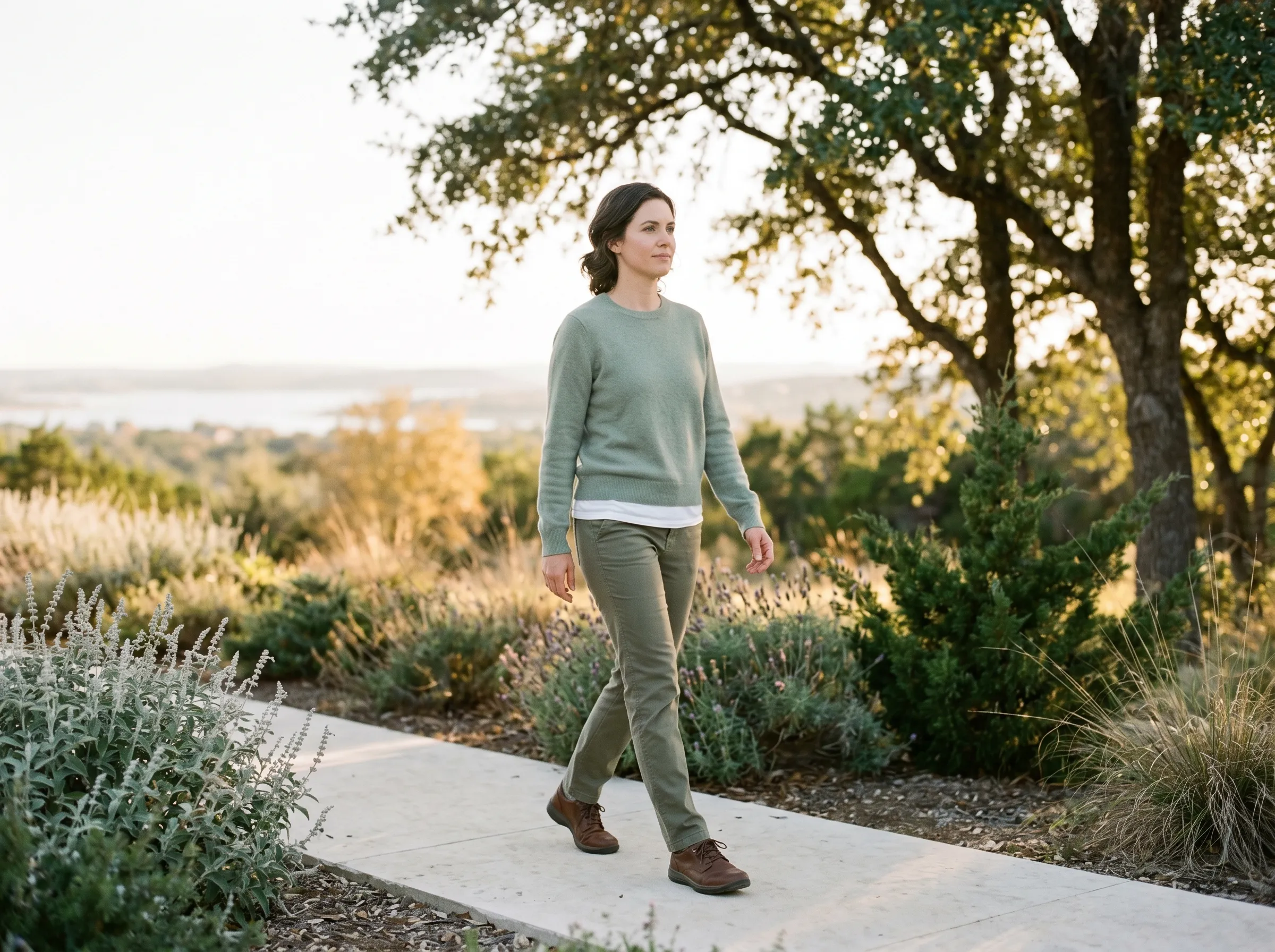 White woman in her early thirties walking forward on a smooth limestone Hill Country path edged with native silver salvia and lavender, live-oak canopy overhead and distant Lake Travis horizon in soft golden hour bokeh.