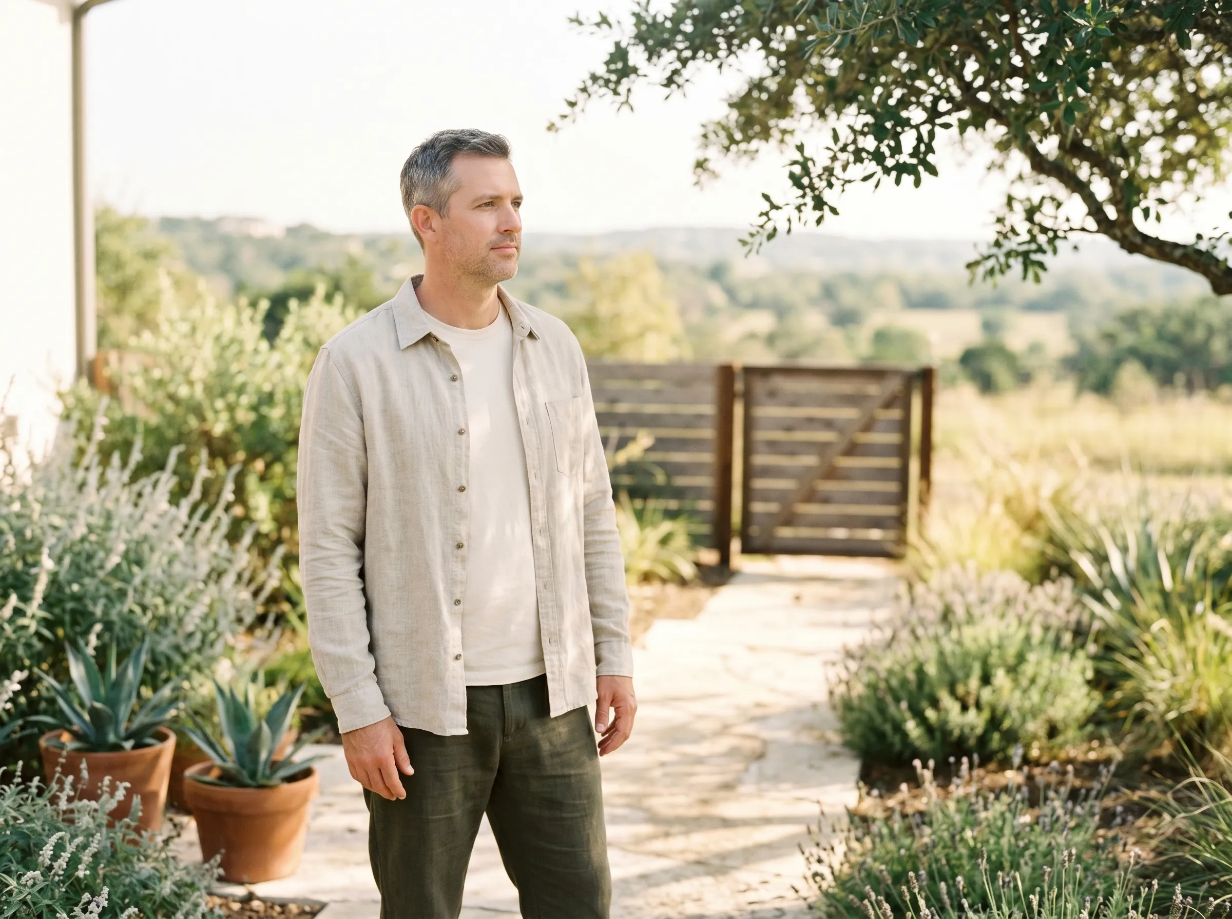 A white man in his mid-40s standing three-quarter profile on a sun-warmed limestone garden path in a Hill Country contemporary side yard, hands loosely at his sides, calm grounded gaze on the middle distance, native silver salvia and agave in terracotta pots flanking the path, weathered cedar gate behind, live-oak canopy softening the warm late-morning light from above.