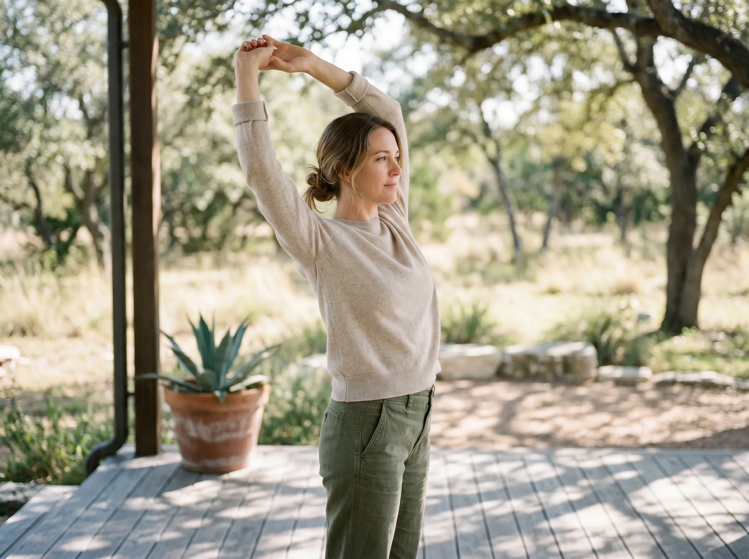 A woman gently stretching her arms overhead on a sunlit Hill Country deck, agave in terracotta nearby, dappled live-oak canopy above.