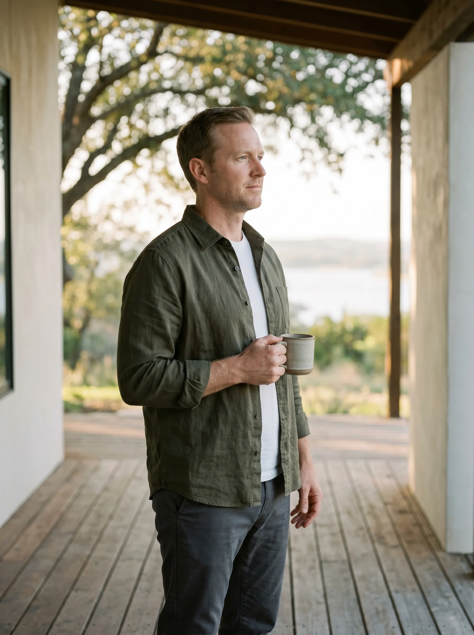 A man in three-quarter profile on a covered Hill Country porch at golden hour, holding a stoneware mug at chest height with Lake Travis softly visible in the middle distance.