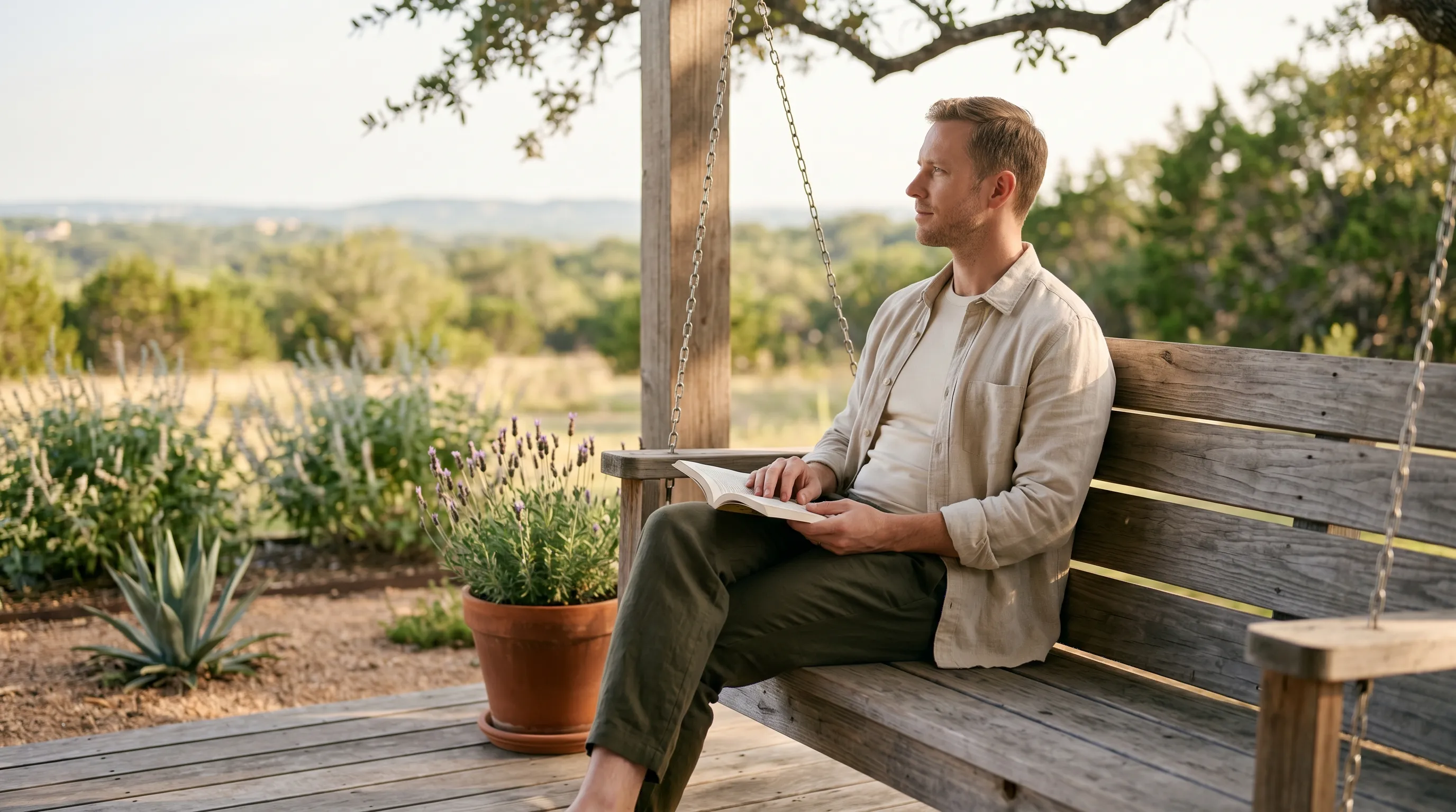 A white man in his mid-30s seated three-quarter profile on a wide weathered cedar plank back-porch swing-bench in a Hill Country backyard, one knee crossed casually, holding a small softcover paperback open in his lap, calm reflective gaze on the soft Hill Country horizon, terracotta-potted lavender on the deck nearby, live-oak canopy softening the late golden afternoon light from above.